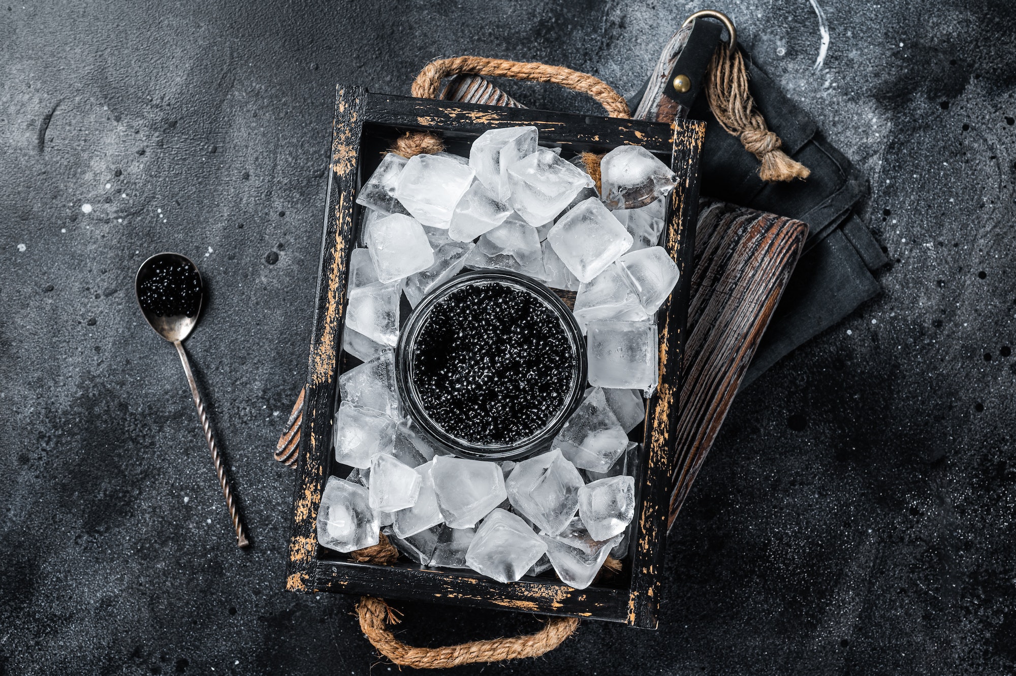 Black caviar served with ice in glass bowl, spoon with black caviar. Black background. Top view